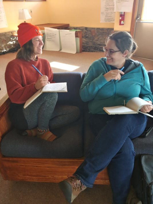 Kateri Boucher and Lydia Wylie-Kellermann sit on a couch with open notebooks, talking during a staff retreat.
